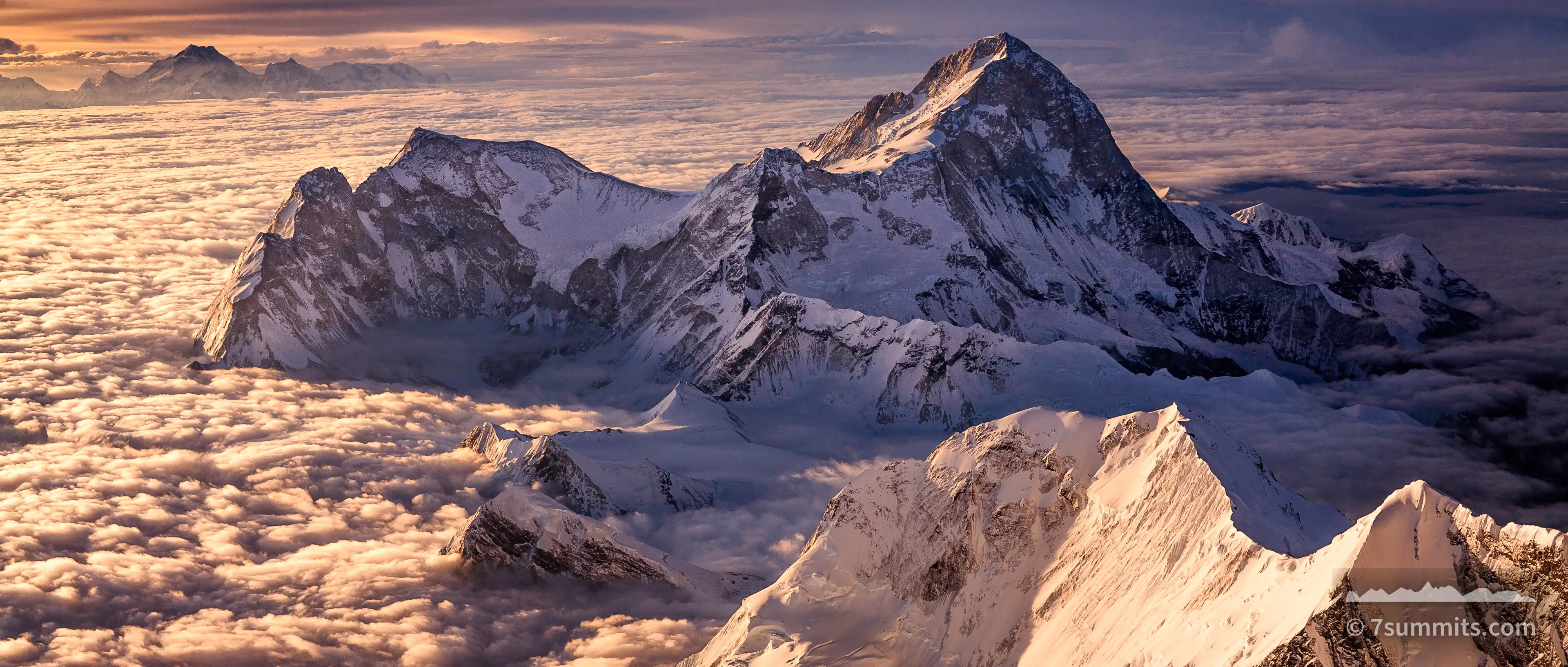 Snow and rock summits rise above an unbroken sea of cloud, lit low and warm from the side, under a streaked violet and orange sky.